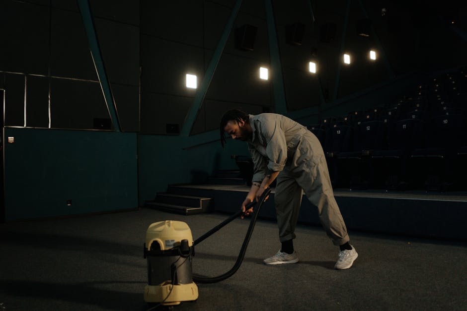 A person dressed in orange work trousers and grey shoes is using a yellow and black vacuum cleaner with a long hose and flat nozzle to perform surface cleaning on a tile floor in a modern living room.