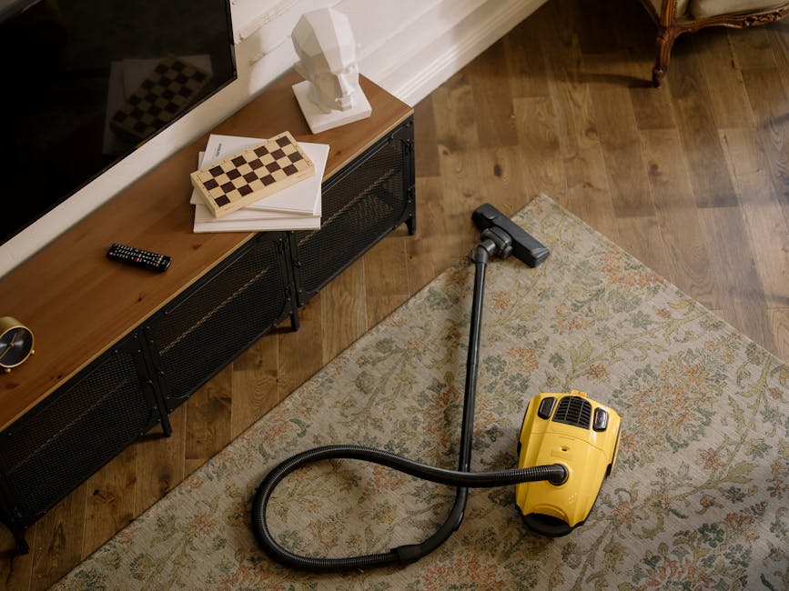 Close-up view of a modern yellow and black vacuum cleaner with a flexible hose attachment on a patterned area rug in a living room. The room features hardwood flooring with a natural finish, a low black metal console table against a white wall, and decorative items including a white bust sculpture, a chessboard, and a remote control. The scene is well-lit with natural light, highlighting the tidy and clean appearance of the surface. This image demonstrates professional deep carpet cleaning and surface maintenance by Enfield Cleaner, focused on improving hygiene and appearance through thorough cleaning processes for domestic spaces.