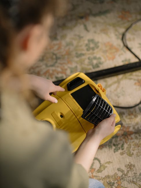 A professional cleaner from Enfield Cleaner dressed in a white protective suit, gloves, and boots is performing deep carpet cleaning in a bright, minimalist living room in Ponders End. The cleaner is using a vacuum or extraction machine with a hose attachment to thoroughly clean a beige carpet, which appears to be wet and fluffy from the cleaning process. The room features natural light coming through a large window, modern decor including potted plants, a wooden sideboard, and a small table, all contributing to a clean and tidy environment. Visible items such as a whiteboard and a wire grid adorn the white walls, emphasizing a neat, organized space. This surface cleaning process demonstrates [COMPANY_NAME]'s expertise in deep cleaning and sanitisation of domestic carpets to ensure hygiene and freshness.