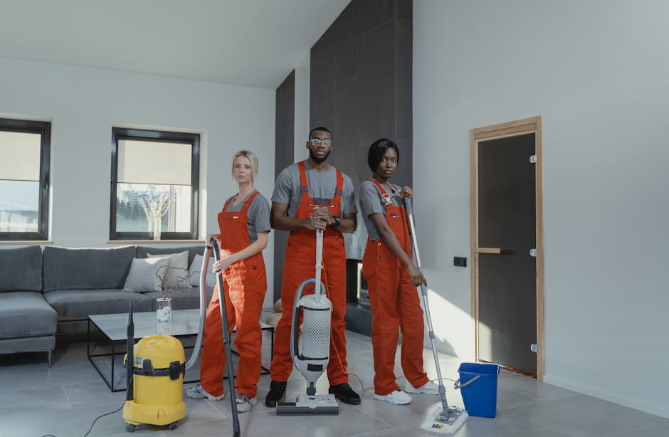 Three professional cleaners dressed in grey t-shirts and red overalls stand in a modern living room during a cleaning session. The room features a black sofa with decorative pillows, a glass coffee table, and large windows allowing natural daylight. They are equipped with various cleaning tools: one holds a vacuum cleaner, another has a mop, and the third has a squeegee, with a yellow carpet extractor and a blue bucket nearby. The surfaces—wooden flooring, glass windows, and a smooth painted wall—appear clean and well-maintained, reflecting a thorough surface cleaning and sanitisation process. The room's minimalist decor and bright lighting highlight the cleanliness efforts, with the professional team from Enfield Cleaner visibly engaged in domestic cleaning tasks consistent with the house cleaning guide for Enfield Town and EN1.