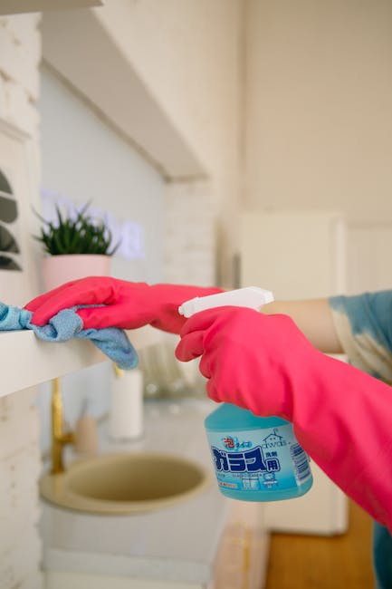 Close-up of a person wearing pink rubber gloves, using a spray bottle of cleaning product to sanitize a white shelf in a residential kitchen. The shelf has a small potted plant and a cloth placed on it, with a stainless steel sink and a white brick wall in the background. The scene is well-lit, emphasizing the cleanliness and hygiene of the surface, highlighting the process of domestic surface cleaning by Enfield Cleaner, a specialist in home and commercial cleaning services in Enfield Town and EN1.