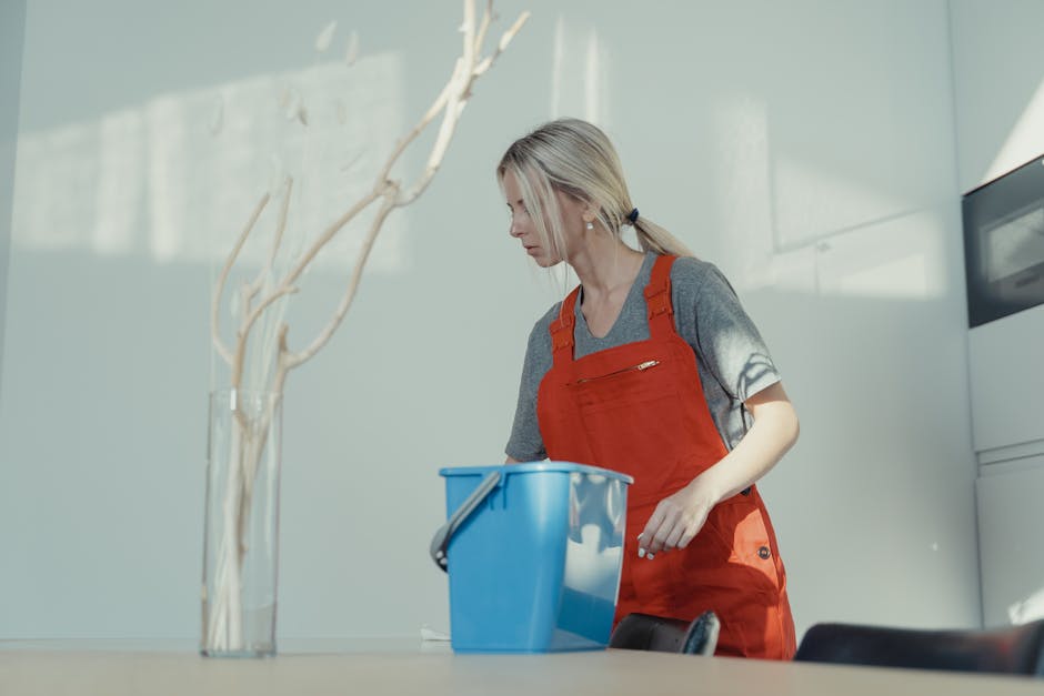 Three professional cleaners dressed in grey t-shirts and red overalls stand in a modern living room during a cleaning session. The room features a black sofa with decorative pillows, a glass coffee table, and large windows allowing natural daylight. They are equipped with various cleaning tools: one holds a vacuum cleaner, another has a mop, and the third has a squeegee, with a yellow carpet extractor and a blue bucket nearby. The surfaces—wooden flooring, glass windows, and a smooth painted wall—appear clean and well-maintained, reflecting a thorough surface cleaning and sanitisation process. The room's minimalist decor and bright lighting highlight the cleanliness efforts, with the professional team from Enfield Cleaner visibly engaged in domestic cleaning tasks consistent with the house cleaning guide for Enfield Town and EN1.