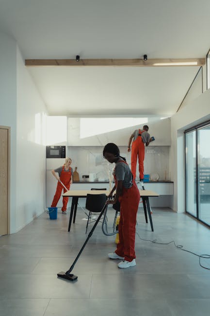 A team of three professional cleaners are performing surface cleaning and deep cleaning in a modern kitchen area with large glass sliding doors, white walls, and grey tiled flooring. One cleaner is vacuuming the light grey tiled floor with a handheld vacuum cleaner, while another is wiping the kitchen countertop near the stove with a cloth. The third cleaner is standing on a step ladder, cleaning the upper part of the wall or cabinet area. The kitchen features minimalist design elements with a wooden beam across the ceiling, stainless steel appliances, and natural light flooding in through the glass doors. The scene highlights thorough sanitisation and detailed cleaning processes, demonstrating the expertise of Enfield Cleaner in domestic cleaning, end of tenancy cleaning, and surface maintenance.