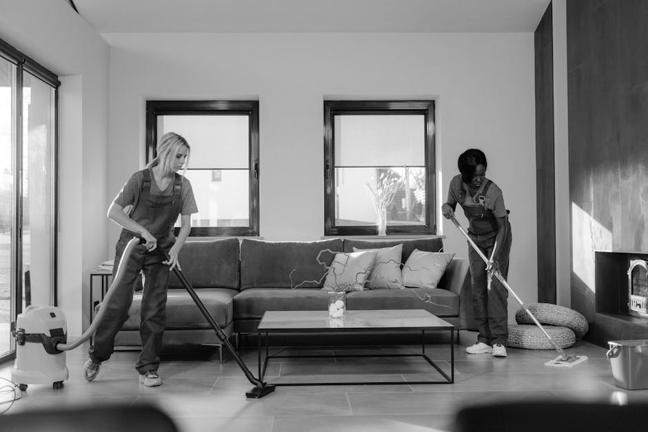 Two professional cleaners from Enfield Cleaner are performing surface cleaning in a modern living room. They are using vacuum and floor mops to thoroughly clean the wooden flooring and the area around a contemporary sofa with cushions. The room features large windows with natural light, a glass coffee table, and minimal decor. The cleaners are wearing grey uniforms with aprons, focusing on maintaining hygiene and a spotless environment, consistent with same-day end of tenancy cleaning services offered by Enfield Cleaner. The scene depicts attention to detail in deep cleaning and sanitisation of residential living spaces.