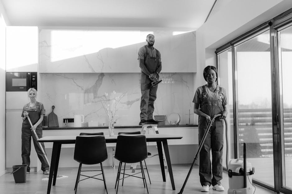 A team of three professional cleaners are performing surface cleaning and deep cleaning in a modern kitchen area with large glass sliding doors, white walls, and grey tiled flooring. One cleaner is vacuuming the light grey tiled floor with a handheld vacuum cleaner, while another is wiping the kitchen countertop near the stove with a cloth. The third cleaner is standing on a step ladder, cleaning the upper part of the wall or cabinet area. The kitchen features minimalist design elements with a wooden beam across the ceiling, stainless steel appliances, and natural light flooding in through the glass doors. The scene highlights thorough sanitisation and detailed cleaning processes, demonstrating the expertise of Enfield Cleaner in domestic cleaning, end of tenancy cleaning, and surface maintenance.